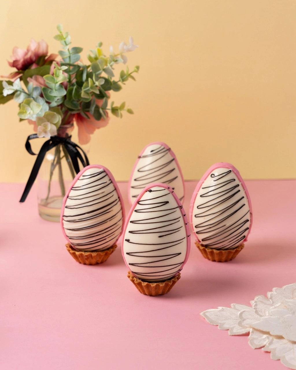 easter white chocolate eggs with pink shell and dark chocolate drizzle design on pastel pink surface with yellow background, premium festive desserts from red ribbon cakes & bistro dubai