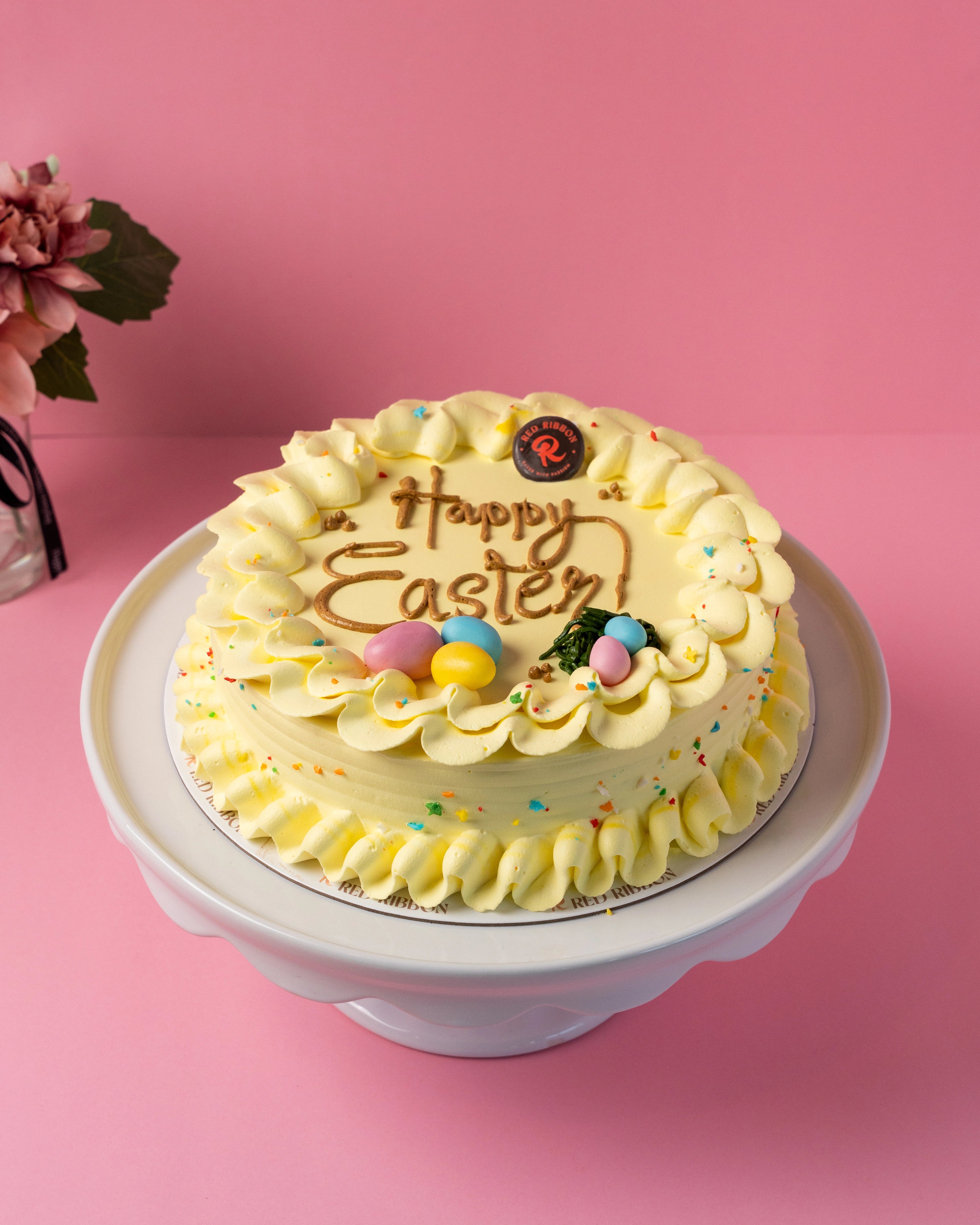 easter celebration cake with happy easter message, creamy frosting, pastel eggs and festive decorations on pink background, red ribbon cakes & bistro dubai