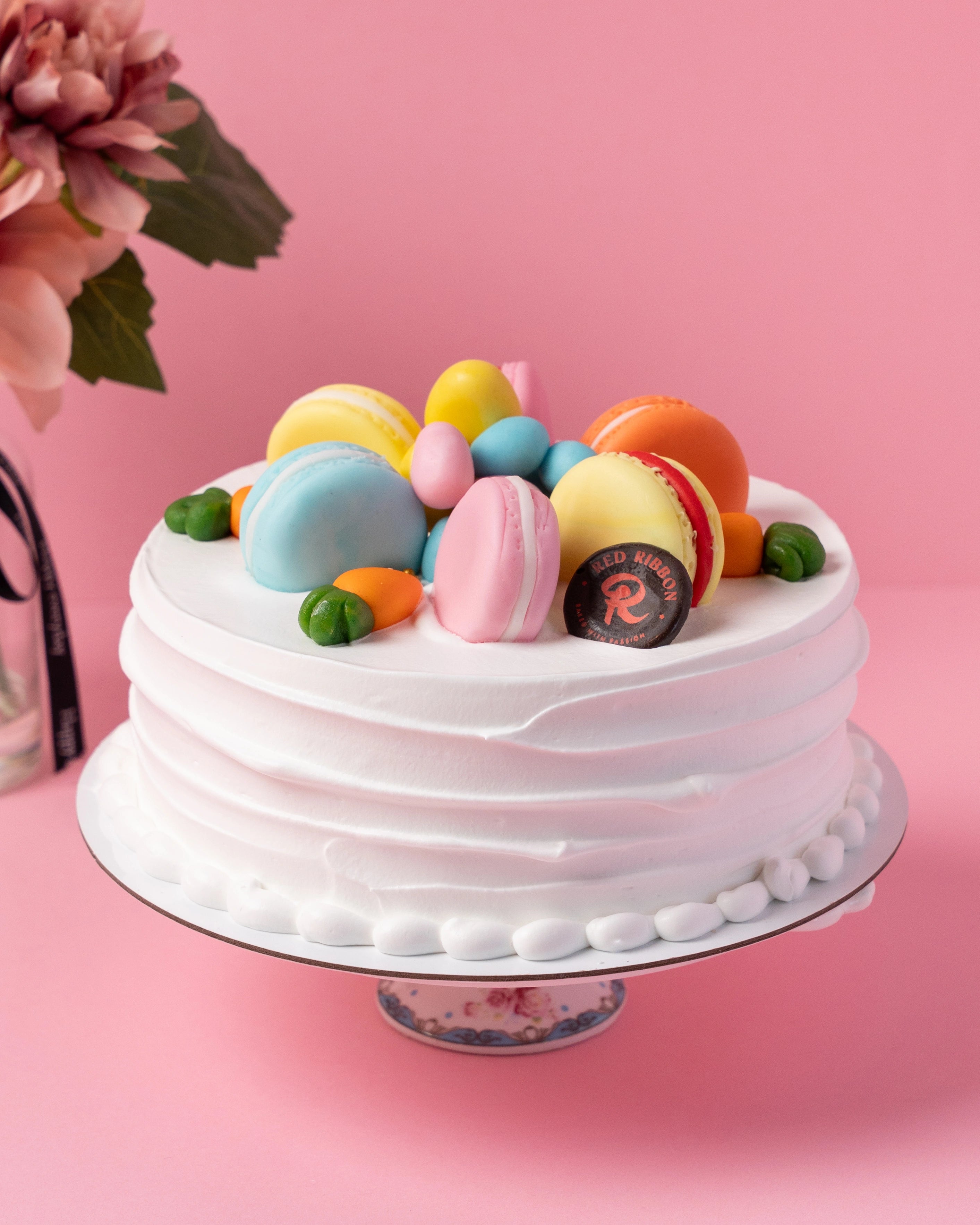 easter themed white frosted cake with colorful macarons and chocolate eggs on pink background, red ribbon cakes dubai