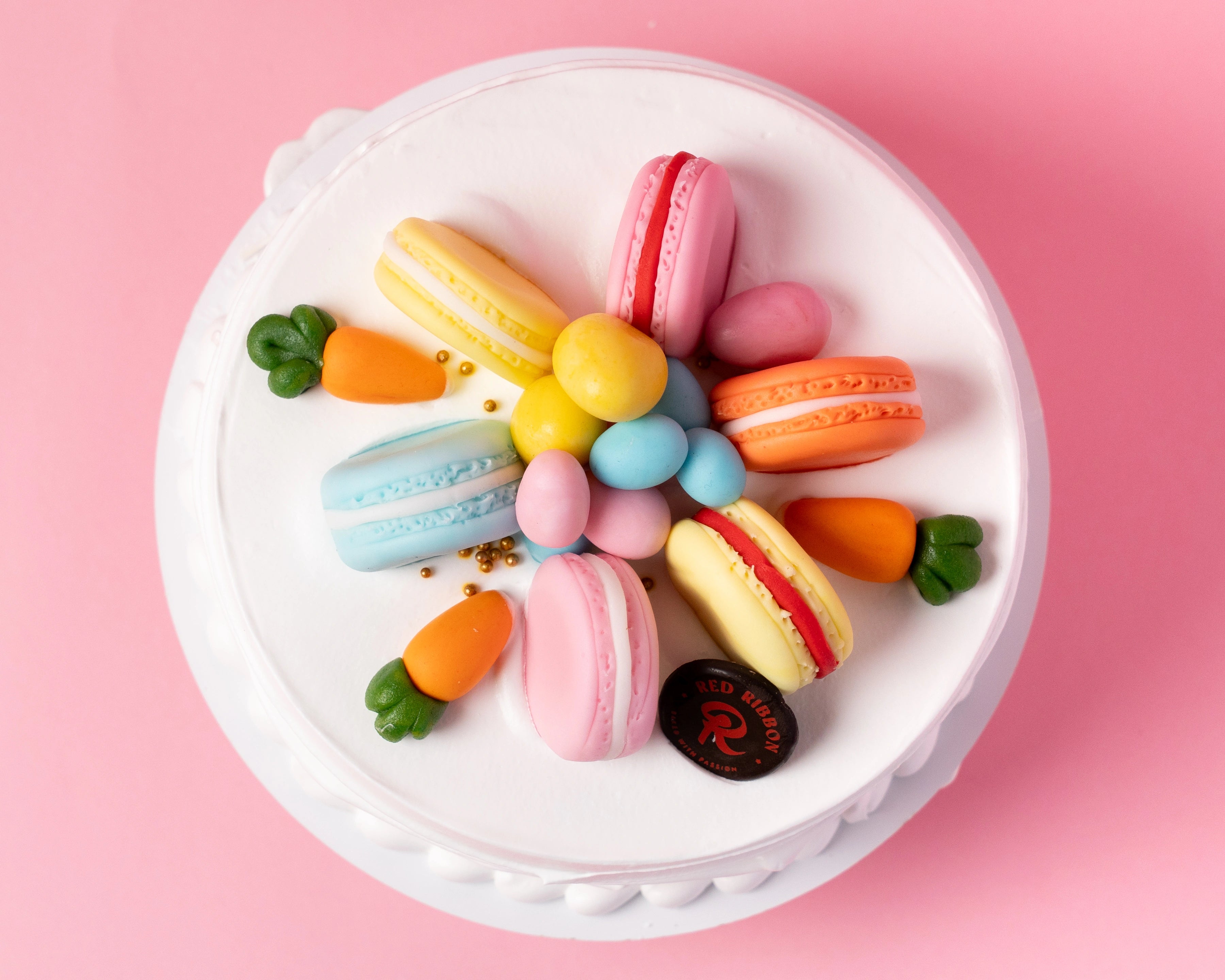 easter celebration cake with smooth white cream frosting topped with pastel macarons and decorative chocolate eggs on soft pink backdrop, red ribbon cakes & bistro dubai